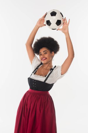 fröhliche afrikanisch-amerikanische Kellnerin im Dirndl mit Fußballball in erhobenen Händen auf weiß