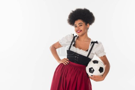 african american oktoberfest waitress in bavarian costume with soccer ball and hand on hip on white