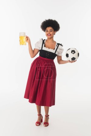 afrikanisch-amerikanische Bayerische Kellnerin posiert mit Fußball und einem Becher Craft Beer mit Schaum auf Weiß