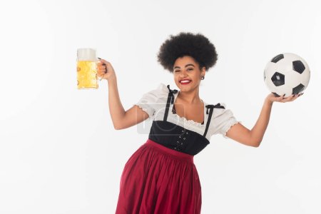 fröhliche afrikanisch-amerikanische Oktoberfestkellnerin mit Krug Bier mit Schaum und Fußball auf weiß