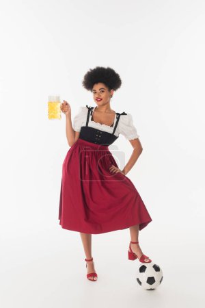 happy african american bavarian waitress posing with mug of beer with foam near soccer ball on white