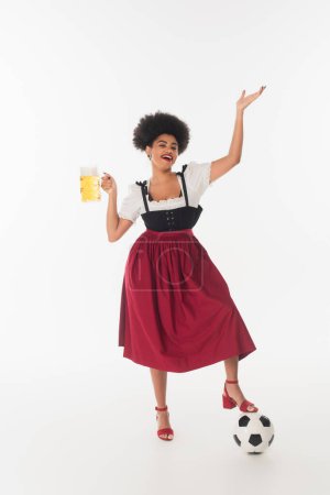 alegre afroamericana oktoberfest camarera con taza de cerveza espumosa artesanal cerca de pelota de fútbol en blanco