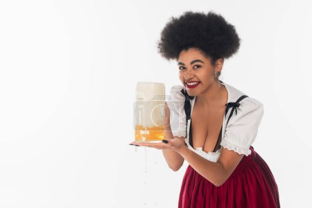 african american oktoberfest waitress holding mug of beer with foam and looking at camera on white