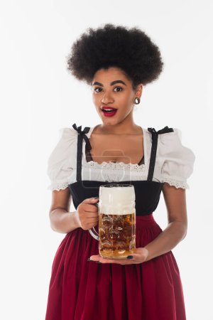 amazed african american bavarian waitress in oktoberfest costume with mug of foamy beer on white