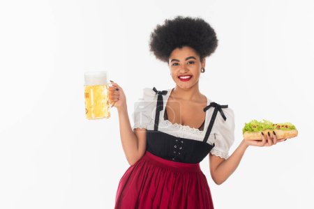 gaie serveuse bavaroise afro-américaine avec tasse de bière et hot dog sur blanc, concept oktoberfest