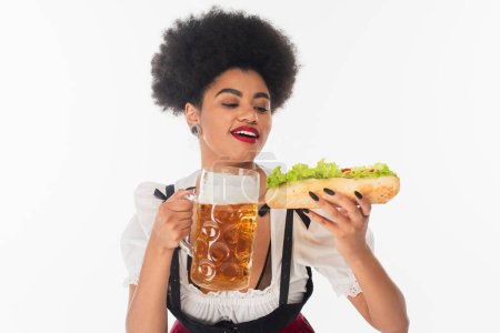 serveuse bavaroise afro-américaine souriante avec tasse de bière et hot dog savoureux sur blanc, oktoberfest