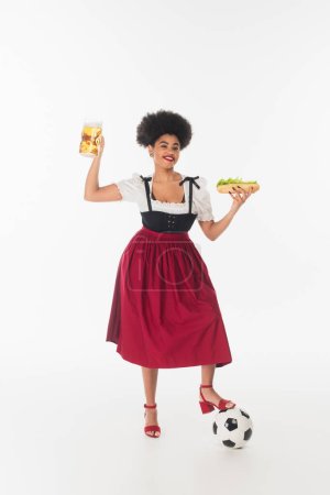 happy african american bavarian waitress in dirndl with beer mug, hot dog and soccer ball on white