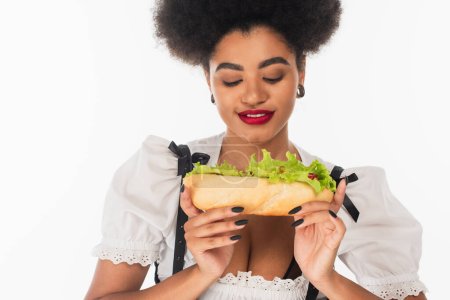 delighted african american oktoberfest waitress in traditional costume with tasty hot dog on white