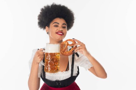 delighted african american oktoberfest waitress with beer mug and pretzel looking at camera on white