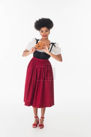 afrikanisch-amerikanische Bayerische Kellnerin im eleganten Dirndl mit leckerer Brezel auf weißem, voller Länge