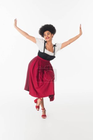 Foto de Alegre africano americano bavarian camarera en elegante dirndl realización oktoberfest danza en blanco - Imagen libre de derechos