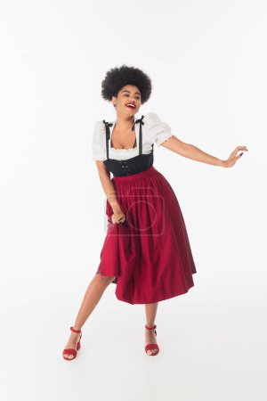energiegeladene afrikanisch-amerikanische Kellnerin in authentischer bayerischer Tracht tanzt beim Oktoberfest auf weiß