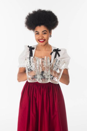 joyful african american bavarian waitress in dirndl holding empty beer mags and smiling on white