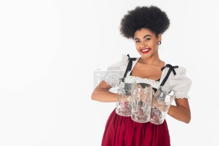 encantadora camarera bavariana afroamericana en traje oktoberfest celebración de revistas de cerveza vacías en blanco