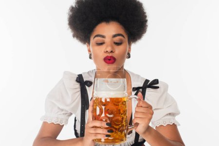 african american bavarian waitress in oktoberfest costume blowing off beer foam in mug on white