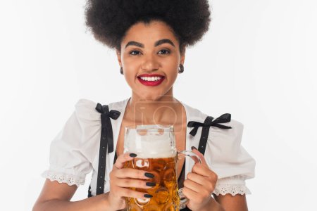 elegant african american oktoberfest waitress with mug of craft beer smiling at camera on white