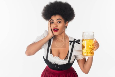 impresionado africano americano oktoberfest camarera sosteniendo taza de cerveza y tocando la cara en blanco