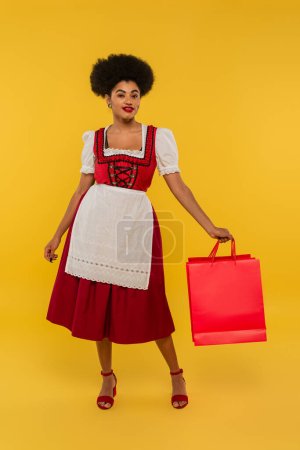 complacida camarera bavariana afroamericana en traje oktoberfest sosteniendo bolsas de compras en amarillo