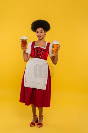 african american waitress in oktoberfest dirndl holding beer mugs and smiling at camera on yellow