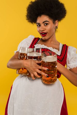 Foto de Emocionada camarera bavariana afroamericana en traje de oktoberfest sosteniendo tres tazas de cerveza en amarillo - Imagen libre de derechos
