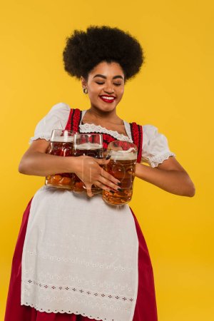 positive african american bavarian waitress in dirndl holding full beer mugs and smiling on yellow