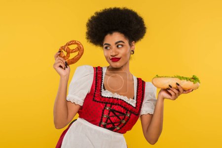 fröhliche afrikanisch-amerikanische Oktoberfestkellnerin in traditioneller Tracht mit Hot Dog und Brezel auf gelb