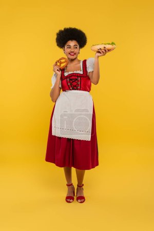 pretty, happy african american bavarian waitress in dirndl posing with hot dog and pretzel on yellow