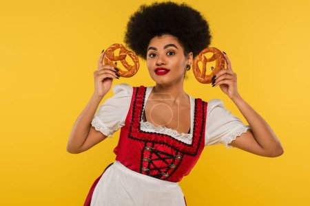 smiling african american oktoberfest waitress in dirndl showing delicious pretzels on yellow