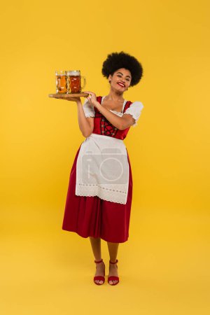 pleased african american bavarian waitress holding wooden tray with beer mugs on yellow, banner