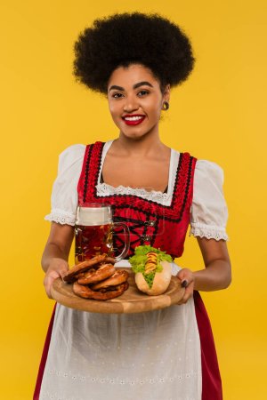 fröhliche afrikanisch-amerikanische Oktoberfestkellnerin mit Holztablett mit Brezeln und Hot Dog auf gelb