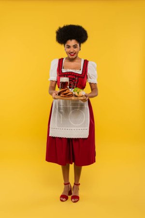 african american oktoberfest waitress holding wooden tray with beer and delicious treat on yellow