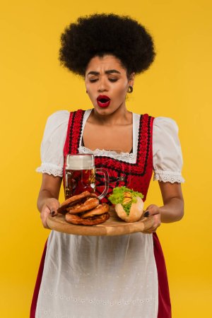 amazed african american bavarian waitress with beer and delicious treat on wooden tray on yellow