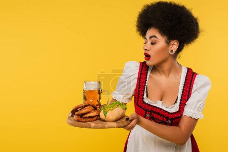excited african american bavarian waitress with beer, hot dog and pretzels on wooden tray on yellow