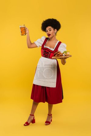 afrikanisch-amerikanische bayrische Kellnerin im Dirndl posiert mit Bier und Essen auf Holztablett auf gelb