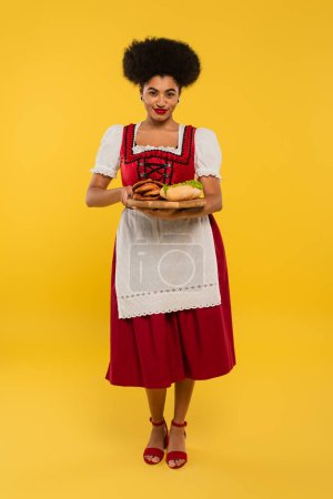 erfreut afrikanisch-amerikanische Bayerische Kellnerin serviert Brezeln und Hot Dog auf Holztablett auf gelb