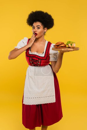 shocked african american bavarian waitress holding wooden tray with pretzels and hot dog on yellow
