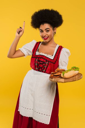 joyful african american oktoberfest waitress with snacks on wooden tray showing idea sign on yellow