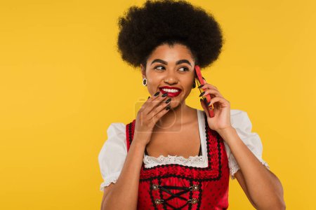 cheerful african american waitress in traditional oktoberfest attire talking on smartphone on yellow
