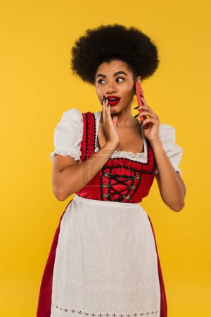 young african american oktoberfest waitress in dirndl costume gossiping on smartphone on yellow