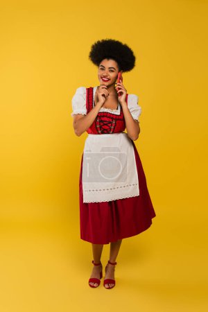 zufriedene afrikanisch-amerikanische Oktoberfestkellnerin im eleganten Dirndl im Handy-Gespräch auf gelb