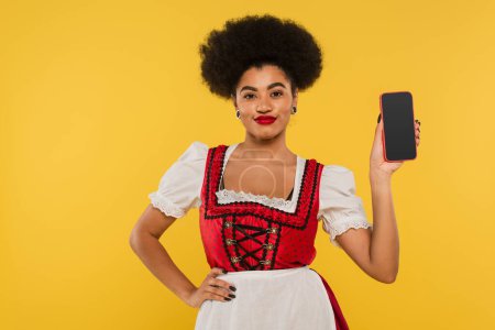 afrikanisch-amerikanische Oktoberfest-Kellnerin im Dirndl zeigt Smartphone mit leerem Bildschirm auf gelb