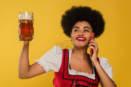 excité africaine américaine oktoberfest serveuse avec tasse de bière parler sur smartphone sur jaune
