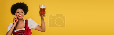 happy african american bavarian waitress with beer mug talking on smartphone on yellow, banner
