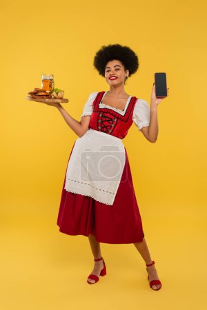 african american bavarian waitress with beer and food showing blank screen of smartphone on yellow