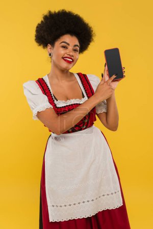 happy african american bavarian waitress in dirndl pointing at blank screen of smartphone on yellow