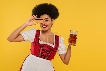 Téléchargez les photos : Gai afro-américain oktoberfest avec tasse de bière visage obscurcissant avec carte de crédit sur jaune - en image libre de droit