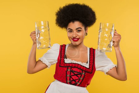 pleased african american oktoberfest waitress in dirndl costume holding empty beer mugs on yellow