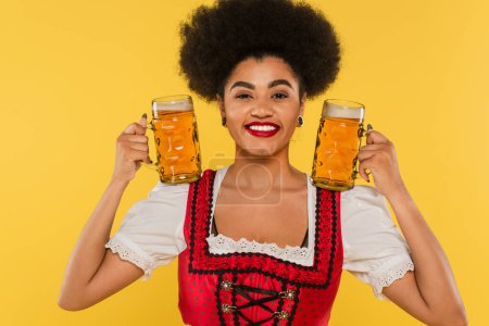 lächelnde afrikanisch-amerikanische Oktoberfestkellnerin im Dirndl mit Bechern mit Craft Beer auf gelb