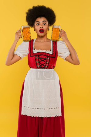 amazed african american waitress in traditional bavarian costume holding beer mugs on yellow