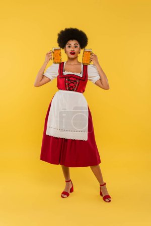 Foto de Impresionada camarera afroamericana en traje bavariano tradicional sosteniendo tazas de cerveza en amarillo - Imagen libre de derechos